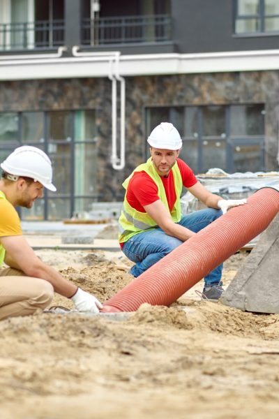 Working mood. Two men in protective helmet and bright vest crouched touching pipe on sand at construction site during day